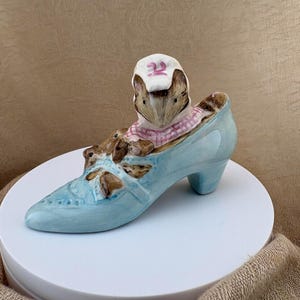 May include: A ceramic figurine of a brown mouse wearing a white bonnet with a pink number 22, sitting inside a light blue high-heeled shoe. The shoe features a pointed toe, a small heel, and decorative brown accents on the top surface.