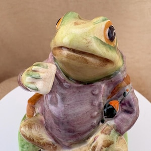 May include: A ceramic frog figurine wearing a purple jacket, seated on a green base. The frog has green skin with brown markings, large dark eyes with orange accents, and is holding a small object in one hand while resting on a green surface.