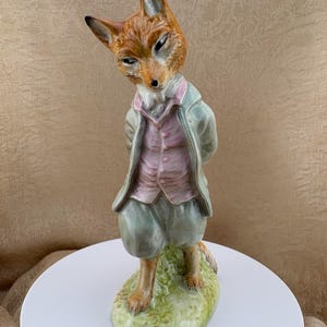 May include: A porcelain figurine of an anthropomorphic fox standing on a green base. The fox wears a light green jacket, a pale pink buttoned vest, and matching light green breeches. The fox has orange and white fur, pointed ears, and a tilted head.