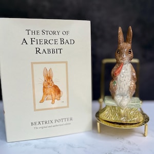 Puede incluir: Un libro titulado The Story of a Fierce Bad Rabbit de Beatrix Potter junto a una figura de conejo de cerámica sentada sobre una base verde, colocada en un pequeño soporte de metal dorado. La portada del libro muestra una ilustración de conejo.