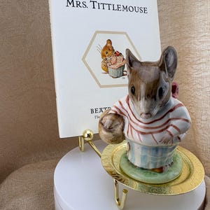 May include: A ceramic figurine of a mouse wearing a red and white striped shirt and a blue apron, standing on a small gold-colored plate. Behind the figurine is a white book titled The Tale of Mrs. Tittlemouse with a small illustration of a mouse.