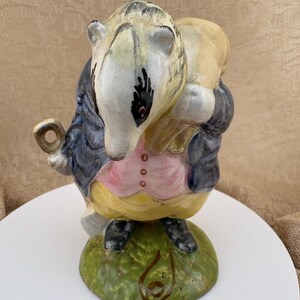 May include: A ceramic figurine of a badger wearing a blue jacket, a pink vest with three buttons, and yellow trousers. The badger stands on a textured green base and carries a light-colored sack over its shoulder, with a small loop held in one hand.