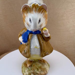 May include: A ceramic figurine of a mouse dressed in a brown coat, a blue scarf, and yellow trousers. The mouse has white and brown fur, pointed ears, and is holding a small yellow object in one hand. It stands on a textured, earthy-colored base.