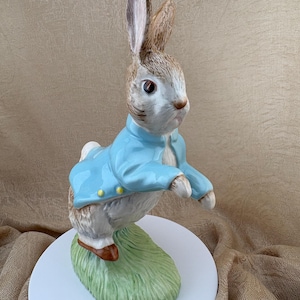 May include: A ceramic figurine of a rabbit standing on a green grassy base. The rabbit has brown and white fur, dark eyes, and is wearing a light blue jacket with small yellow buttons. The figure is depicted in a standing, upright position.