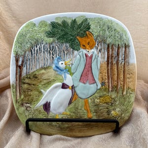 May include: A square ceramic decorative plate featuring a raised relief illustration of a fox wearing a light blue coat and red vest walking alongside a white duck wearing a blue bonnet and purple sash in a forest setting with green trees.