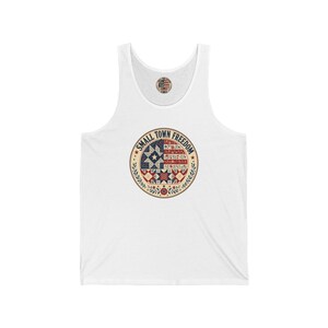Peut inclure: Un débardeur blanc avec un motif circulaire sur la poitrine. Le design comprend le texte 'SMALL TOWN FREEDOM' entourant un motif inspiré du patchwork en rouge, blanc et bleu, avec des accents floraux et une étoile au centre.