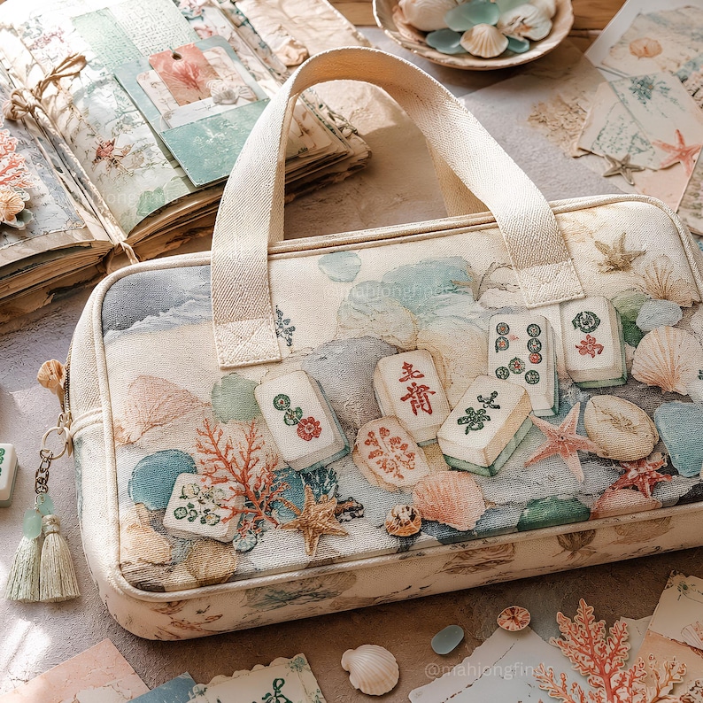 Op de afbeelding: Een rechthoekige, cr&egrave;mekleurige stoffen tas met twee handvatten, voorzien van een print met verspreide mahjongtegels, schelpen, zeesterren en koraal. Een kleine decoratieve kwast met lichtgroene kralen hangt aan de rits aan de zijkant.