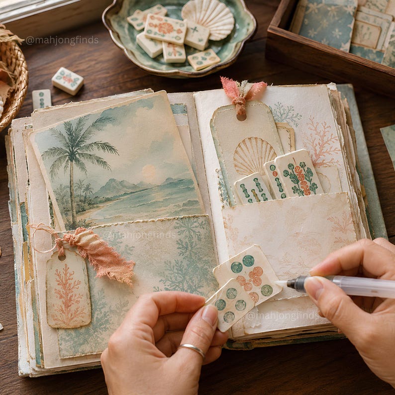 Op de afbeelding: Een open plakboek met pagina's in vintage-stijl, voorzien van een aquarelillustratie van een tropisch strand, papier met koraalpatronen en decoratieve labels. Kleine rechthoekige mahjongtegels met groene en oranje ontwerpen zitten in de vakjes.