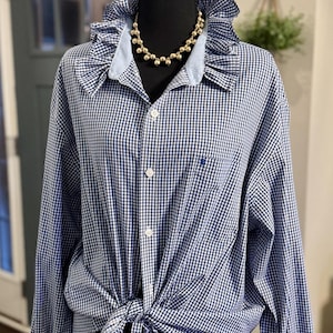 May include: A blue and white gingham shirt with a ruffled collar and light blue cuffs. The long-sleeved shirt is tied at the waist and has a small embroidered logo on the chest. A gold beaded necklace is worn with the shirt.