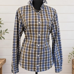 May include: A long-sleeve button-down shirt featuring a plaid pattern in shades of olive green, navy blue, and white. The shirt has a standard collar, a front button placket with white buttons, and barrel cuffs on the sleeves, displayed on a mannequin.