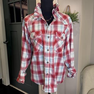 May include: A long-sleeve button-down shirt featuring a red, white, and light blue plaid pattern. The shirt has a ruffled collar, two chest pockets with button closures, and a central button placket. The Wrangler brand logo is visible on the pocket.