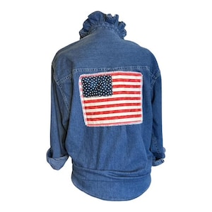 May include: A blue denim jacket featuring a ruffled collar and long sleeves with rolled cuffs. The back of the jacket displays a rectangular patch of the American flag, characterized by a blue field with white stars and horizontal red and white stripes.