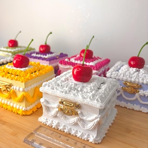 May include: A selection of square decorative boxes, each topped with a bright red cherry. The boxes are decorated with frosting-style detailing in colours such as yellow, white, purple, and pink. Gold-coloured clasps secure the lids. A ruler is placed in front of the boxes.