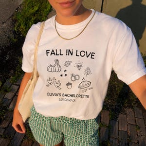 May include: A white t-shirt featuring black line art graphics including pumpkins, leaves, a lightbulb, two rings, two birds, an acorn, and a pie. The text reads FALL IN LOVE, OLIVIA'S BACHELORETTE, SAN DIEGO CA, and EST. 2026. A gold chain necklace is worn.