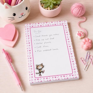 Può includere: Un blocco note rettangolare con centro bianco e bordo rosa decorato con piccoli cuori e impronte di zampe. Il testo recita To-Do: Send thank you notes, Pick up cat food, Water plants, Call Mom, Plan weekend lunch. In basso è raffigurato un gatto.