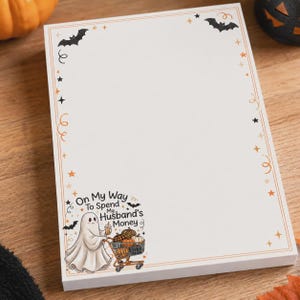 May include: A rectangular notepad with a white background and an orange border. The border features black bats, orange stars, and swirls. In the bottom left corner, a ghost pushes a shopping cart filled with pumpkins, with the text On My Way To Spend My Husband's Money.