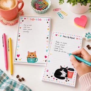 Puede incluir: Dos blocs de notas rectangulares de papel con ilustraciones de gatos. Uno se titula Coffee & Cats y muestra un gato atigrado naranja con una taza. El otro se titula Paws, Sip, Repeat y muestra un gato blanco y negro junto a un vaso de café.
