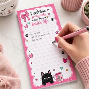 Può includere: Un blocco note rettangolare rosa e bianco con bordo smerlato, caratterizzato dal testo I work hard so my cats can have a better life, un'illustrazione di gatto nero, una tazza rosa con la scritta Cat Mom Fuel, impronte di zampe e cuori.