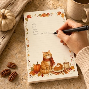 Puede incluir: Un bloc de notas rectangular con papel rayado y un borde con temática otoñal de hojas, bayas y calabazas. La página incluye una ilustración de un gato atigrado naranja con una bufanda roja, un trozo de tarta de calabaza y una taza.