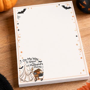 May include: A rectangular notepad with a white background and a decorative border featuring black bats, orange stars, and swirls. The bottom left corner displays a ghost pushing a shopping cart filled with pumpkins and the text On My Way To Spend My Husband's Money.