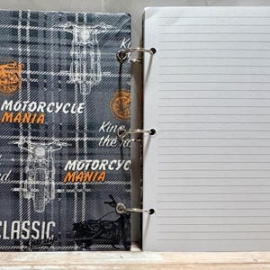 May include: A spiral-bound notebook featuring a dark gray plaid cover with white and orange motorcycle illustrations. The cover displays the text Motorcycle Mania, King of the road, and Classic Garage. The notebook has white lined paper pages.