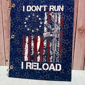 May include: A blue notebook with a repeating star pattern featuring a distressed American flag graphic, a rifle illustration, and the white text I DON'T RUN I RELOAD. The cover is secured with three silver metal binder rings on the left side.