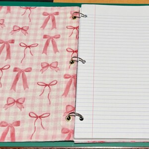 May include: A three-ring binder featuring a fabric cover with a pink and white gingham pattern decorated with small pink bows. The interior contains white lined paper held in place by three silver metal rings attached to a green spine.