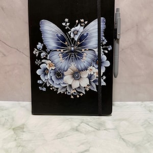 May include: A black hardcover notebook featuring a large blue butterfly surrounded by light blue and white flowers on the front cover. A black elastic band wraps vertically around the notebook, and a grey pen is held in a loop on the right side.