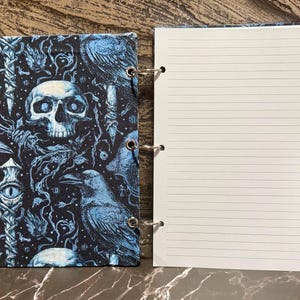 May include: A spiral-bound notebook featuring a black and blue fabric cover with a repeating pattern of skulls, ravens, and mystical eyes. The interior consists of white, lined paper pages held together by three silver metal rings on the side.