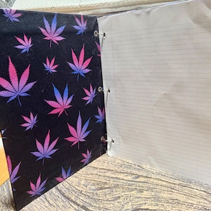 May include: A three-ring binder featuring a black cover with a repeating pattern of pink and blue cannabis leaves. The binder is open, revealing white lined paper held in place by three silver metal rings along the interior spine edge.