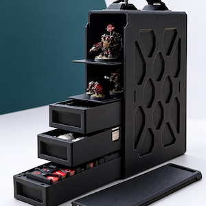 Puede incluir: Un estuche de almacenamiento modular negro con asa superior, que cuenta con estantes abiertos con figuras en miniatura pintadas y tres cajones extraíbles que contienen pequeños componentes de juego y fichas. El panel lateral tiene un patrón hexagonal.