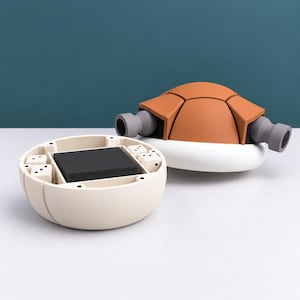 May include: A multi-part game set featuring a cream-coloured circular base with internal compartments holding four white dice and a stack of black cards. Beside it sits a separate orange and white turtle-shaped shell component with grey side ports.