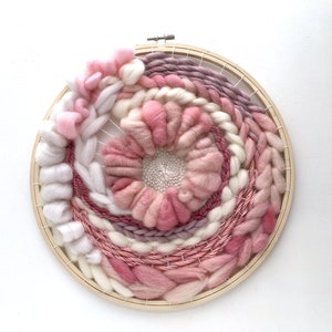 May include: A circular wall hanging woven with pink, white, and purple yarn in a circular pattern. The yarn is woven around a wooden hoop.