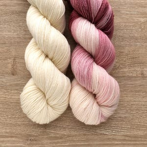 Puede incluir: Dos ovillos de hilo, uno blanco y otro degradado en rosa y rojo.