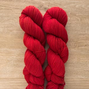May include: Two skeins of vibrant red yarn are displayed against a light wood grain background. The yarn is tightly wound, showcasing its texture and color. This image is ideal for showcasing knitting or crochet supplies.