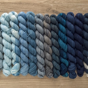 May include: A collection of mini skeins of yarn in a gradient of blue and gray hues. The yarn is arranged in neat rows, transitioning from light blue to deep navy. The skeins are tightly wound, showcasing the texture of the fiber.