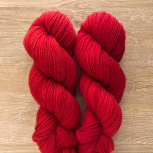 May include: Two skeins of vibrant red yarn are displayed against a light brown wooden surface. The yarn is tightly wound, showcasing its texture and color. This image is ideal for showcasing knitting or crochet supplies.