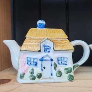 Puede incluir: Una tetera de cerámica con forma de pequeña casa de campo con techo de paja amarillo, exterior blanco, ventanas y puerta pintadas de azul y detalles de follaje verde. La tetera cuenta con un pico vertedor, un asa y una chimenea en la parte superior.