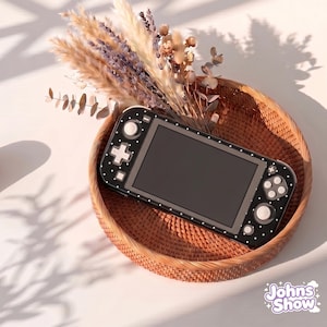 Può includere: Una console Nintendo Switch Lite a pois bianchi e neri è appoggiata in un cestino intrecciato con fiori secchi. La console ha uno schermo nero e pulsanti bianchi. Il cestino è marrone e lo sfondo è bianco.