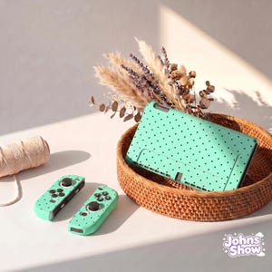 May include: A mint green Nintendo Switch case and controllers with black polka dots are displayed in a woven basket. The controllers and case are on a white surface with a spool of thread and dried flowers.