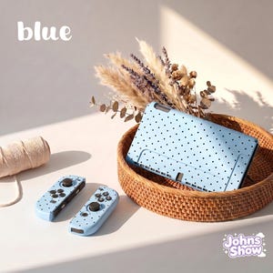 May include: A light blue gaming console and two matching controllers, all featuring a black polka dot pattern. The console is placed inside a round woven basket, while the controllers sit nearby. The word blue is printed in white text.