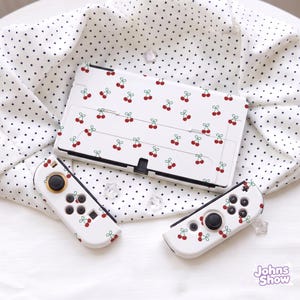 Puede incluir: Una consola de juegos blanca y dos mandos desmontables con un patrón repetitivo de cerezas rojas con tallos verdes. La consola tiene un soporte en la parte posterior. Los mandos incluyen joysticks y botones negros. El texto Johns Show es visible.