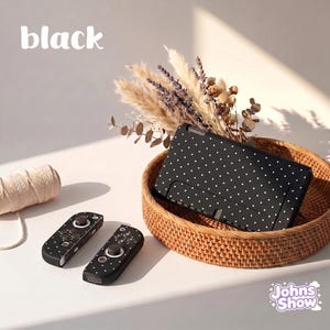 May include: A black video game console and two matching controllers, all featuring a white polka dot pattern. The console is placed inside a round woven basket, and the text "black" and "Johns Show" are visible in the image.
