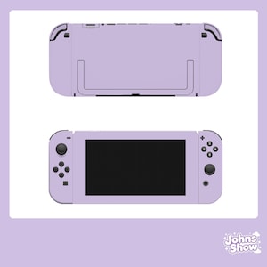 Puede incluir: Una consola de videojuegos de color lila claro, mostrada desde la parte delantera y trasera. La vista frontal muestra una pantalla negra, botones de control y joysticks. La vista trasera muestra un soporte y puertos. La marca "Johns Show" está en la esquina inferior derecha.