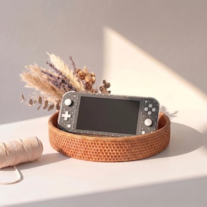 May include: A dark grey handheld gaming console featuring a white dotted pattern and white buttons, resting inside a round woven rattan tray. The console is positioned in front of a small arrangement of dried lavender and beige pampas grass.