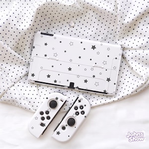 May include: A white gaming console and two matching controllers featuring a black star and dot pattern. The console has a rectangular screen area and a kickstand on the back. The controllers include black joysticks, buttons, and directional pads.