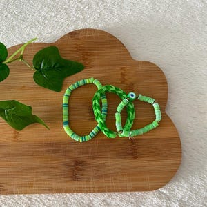 May include: Three bracelets in shades of green. One is a single strand of flat circular beads, one is a braided green cord, and the third is a strand of flat circular beads featuring a small round charm with a blue and white eye design.