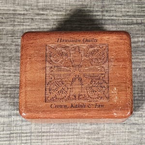 May include: A rectangular wooden box with a medium brown finish. The lid features a laser-engraved square design of a floral pattern. The text "Hawaiian Quilts" is printed above the design, and "Crown, Kahili & Fan" is printed below it.