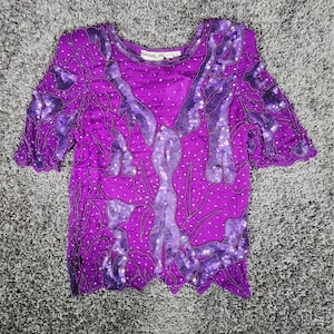 May include: A vibrant purple top with short sleeves, adorned with intricate beadwork and sequin details. The design features darker purple accents and a scalloped hemline. The top is a unique piece of apparel.
