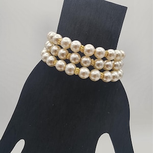 May include: A triple-strand bracelet featuring round, cream-colored faux pearls. Each pearl is separated by small, textured gold-tone metal spacers. The bracelet is arranged in three parallel rows, creating a layered appearance with a consistent design.